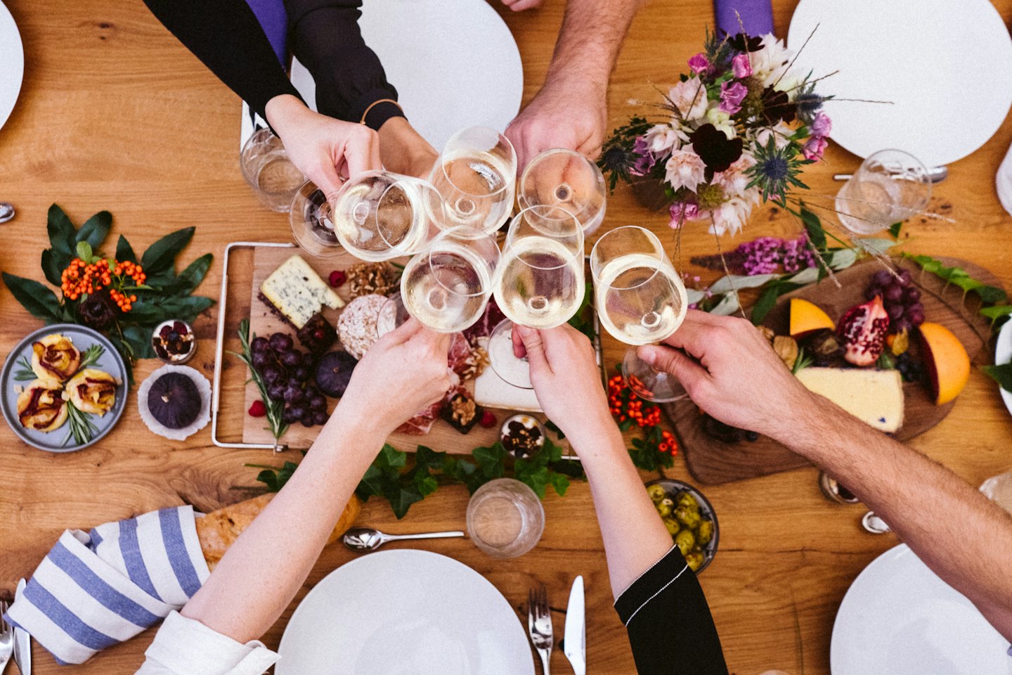 Hands clinking white wine glasses over a table of food.